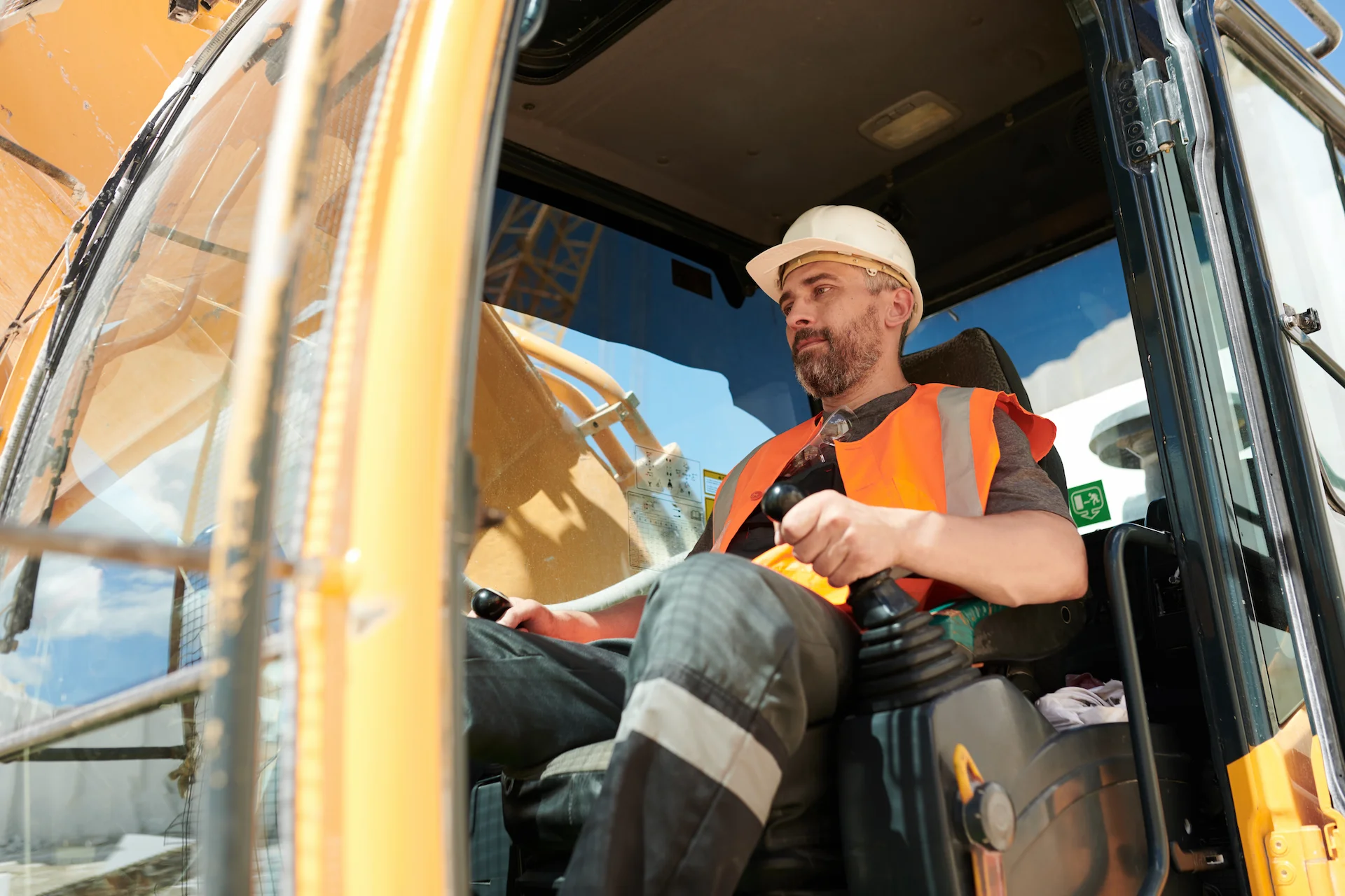 Male builder or driver of construction machine in workwear and hardhat operating caterpillar truck while sitting in cab during work